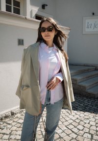 Blazer oversize vert clair sur une chemise rayée rose et blanche, un t-shirt blanc et un jean bleu clair. Des lunettes de soleil foncées complètent le look.