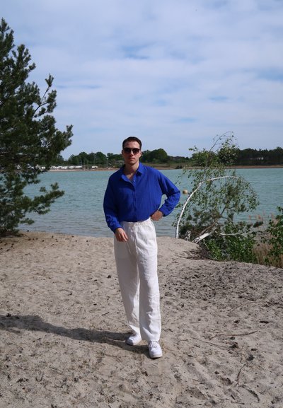 Camisa de manga larga texturizada azul, pantalones de lino blancos y zapatillas blancas. De pie sobre un suelo arenoso cerca de un cuerpo de agua y vegetación.