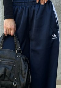 Persoană purtând pantaloni sport Adidas bleumarin, ținând o geantă neagră din piele texturată, cu mânere împletite, pe fundalul unui perete gri.