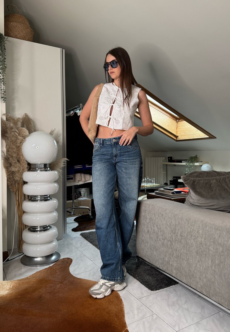 Femme portant des lunettes de soleil, un top court blanc sans manches, un jean bleu et des baskets, debout à l'intérieur près d'un canapé et d'une grande lampe blanche.