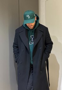 Άνδρας με περιποιημένο μούσι, που φοράει πράσινο hoodie με το γράμμα Α, σκούρο διπλόπατο παλτό και πράσινο καπέλο NY, στέκεται με τα χέρια στις τσέπες, κοιτάζοντας προς τα κάτω.