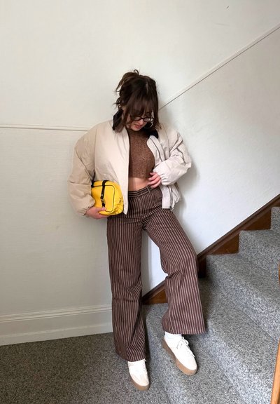 Mujer de pie en escaleras alfombradas, vistiendo una chaqueta acolchada beige, top corto marrón, pantalones a rayas, zapatillas blancas y sosteniendo un bolso pequeño amarillo.