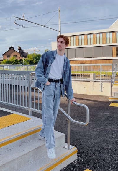Chaqueta de mezclilla azul claro y jeans, camiseta blanca, cinturón negro, de pie junto a una barandilla metálica en las escaleras, con un edificio moderno y vegetación de fondo.