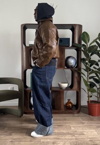 Veste bomber en cuir brun texturé avec une capuche noire, associée à un jean en denim foncé ample et des chaussures blanches avec revers pliés.