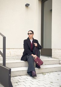 Abrigo negro sobre un suéter burdeos, pantalones anchos grises y un bolso color burdeos. Gafas de sol oscuras y peinado elegante complementan el conjunto.