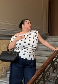 Γυναίκα με μπλούζα πουά και σκούρο παντελόνι, κρατώντας μια μαύρη τσάντα, στέκεται σε εσωτερική πέτρινη σκάλα, κοιτάζοντας αριστερά της.