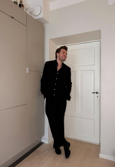 Camisa negra abotonada y pantalones, zapatos brillantes, de pie contra una pared de color claro con una puerta blanca. Gabinetes de cocina modernos visibles.