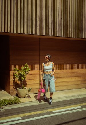 Top corto gris claro, pantalones cortos de denim deslavado y oversize con patrones, bolso rosa, cabello trenzado colorido y zapatillas negras en la calle.