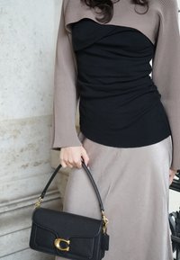 Borsa a mano in pelle nera con hardware dorato, tenuta da una persona che indossa un maglione beige a costine e una gonna liscia di un grigio chiaro.
