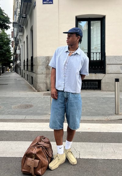 Conjunto casual compuesto por una camiseta de manga corta a rayas de color azul claro, pantalones cortos de mezclilla y zapatillas beiges. Bolsa de viaje marrón en el suelo.