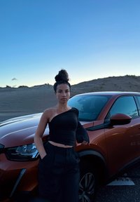 SUV laranja com bordas arredondadas, acabamento brilhante e detalhes em preto. Top cropped preto e calças largas pretas vestidas por uma pessoa encostada no veículo.