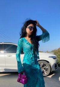 A flowing, translucent turquoise outfit with ruffled accents, paired with a glossy purple handbag and oversized sunglasses.