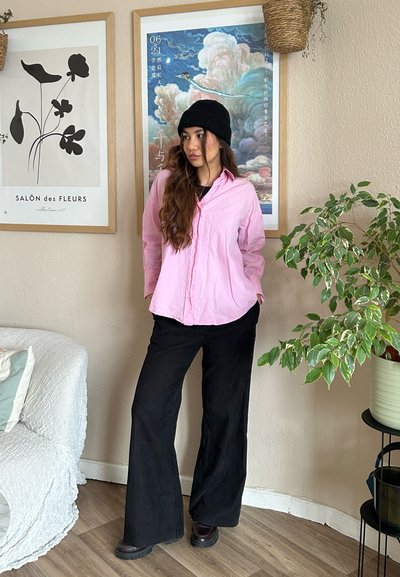 Camisa de color rosa claro, de mangas largas, con cuello abierto y botones en la parte delantera, combinada con pantalones negros de pierna ancha con textura de canalé. Lleva un gorro negro en la cabeza, en un entorno interior.