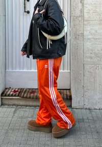 Πορτοκαλί αθλητικά παντελόνια Adidas με τρεις λευκές ρίγες σε κάθε πλευρά, μαύρο δερμάτινο μπουφάν και μπεζ πλατφόρμες. Λευκή τσάντα με φουντάκια.