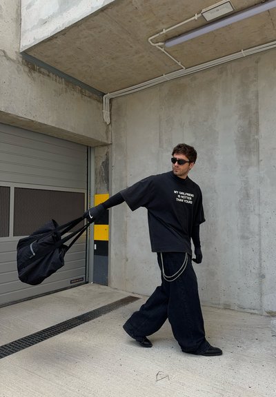 Camiseta negra de gran tamaño con texto, jeans azules holgados, guantes negros y gafas de sol, llevando un bolso negro con correa. Fondo de hormigón.
