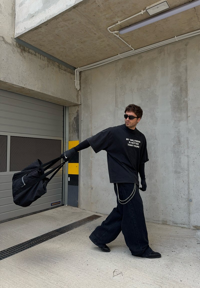 Μαύρη oversized μπλούζα με κείμενο, χαλαρές μπλε τζιν παντελόνες, μαύρα γάντια και γυαλιά ηλίου, κρατώντας μια μαύρη τσάντα με λουρί. Συνοδευτικό φόντο από σκυρόδεμα.