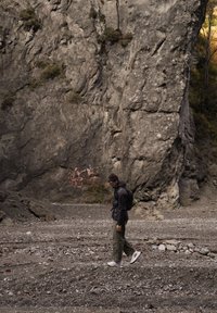 Osoba kráčející po kamínkové zemi u velké skalní stěny, oblečena v tmavé prošívané bundě, hnědé pletené čepici, olivových kalhotách a teniskách.