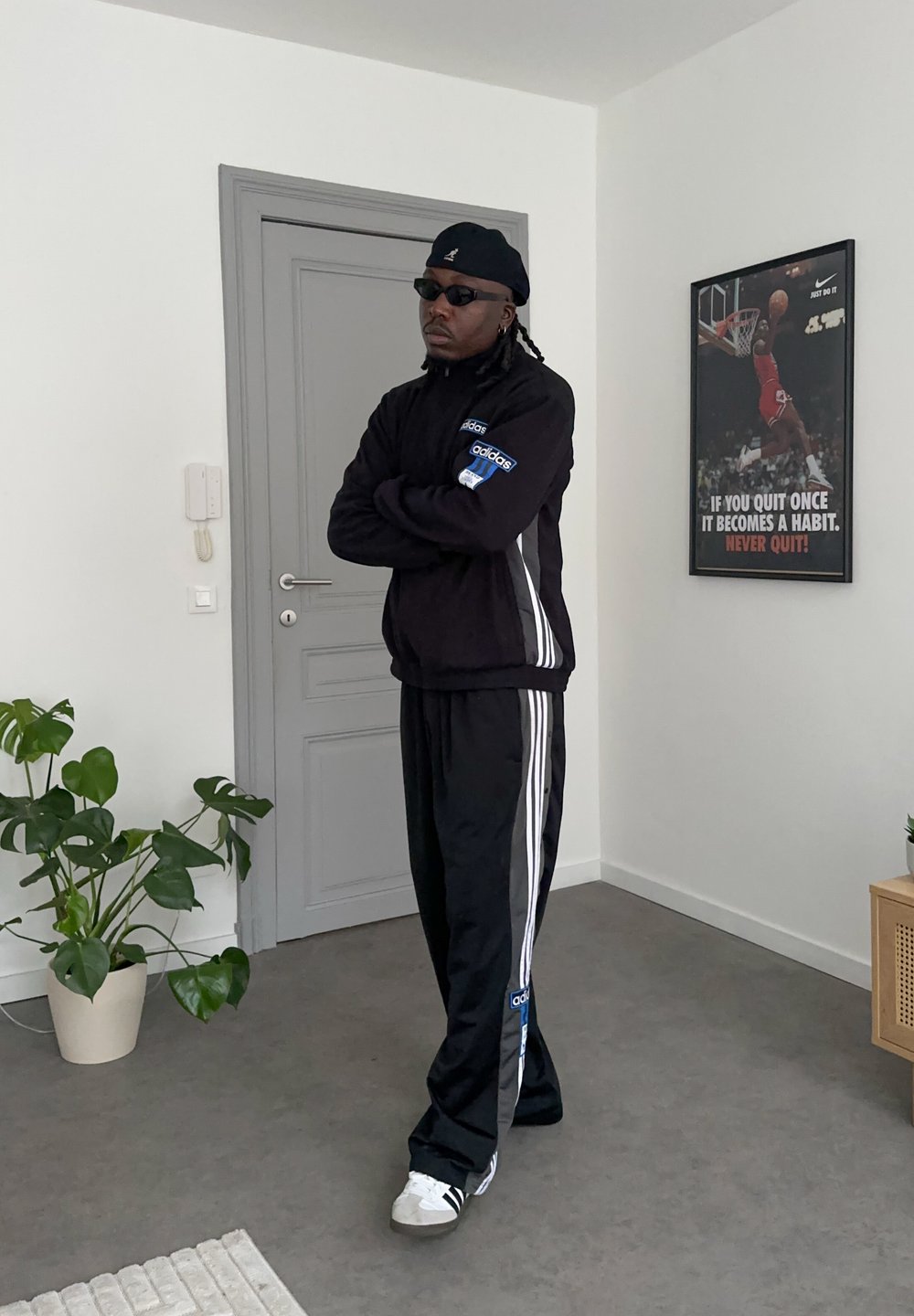 Homme en survêtement Adidas noir, lunettes de soleil noires et chapeau, debout les bras croisés dans une pièce minimaliste avec une affiche de motivation sur le basket-ball.