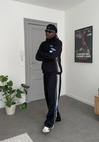 Homme en survêtement Adidas noir, lunettes de soleil noires et chapeau, debout les bras croisés dans une pièce minimaliste avec une affiche de motivation sur le basket-ball.