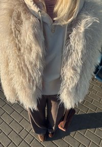 Vestă pufoasă bej deschis peste un hanorac gri, asortată cu pantaloni maro cu croială largă. Detalii suplimentare includ un colier mic și o geantă roz.