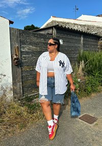 White pinstriped baseball jersey featuring a black logo, distressed denim shorts, red trainers with cream laces, and a blue bag. Set against a backdrop of greenery and a wooden fence.