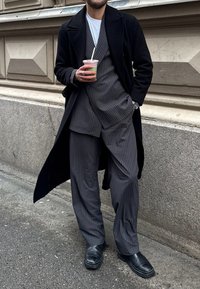 Fato cinza risca de giz com calças de perna larga e um blazer ajustado, sobreposto a um longo casaco preto. Segurando uma bebida, sapatos pretos completam o visual.