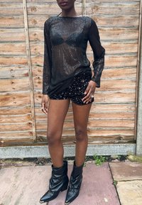 Juoda ilgomis rankovėmis palaidinė su blizgančiomis akutėmis, dėvima virš blizgančių juodų šortų; derinama su juodais atremtais aulo batais. Medinis fonas.