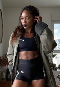 Femme aux cheveux tressés portant un soutien-gorge de sport noir, un short Adidas noir et une veste verte, posant à l'intérieur près d'une fenêtre avec des plantes.