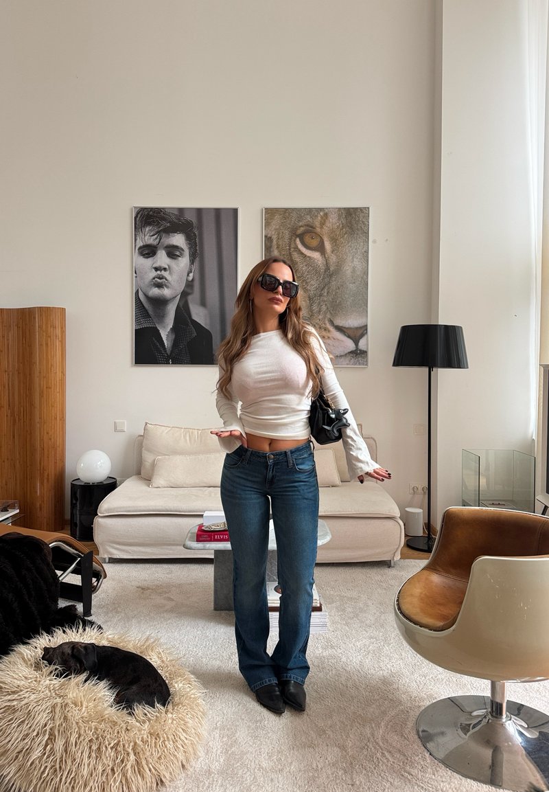Jean flare bleu taille haute, haut blanc à manches longues et lunettes de soleil noires oversize. En arrière-plan, un canapé beige et une œuvre d'art.
