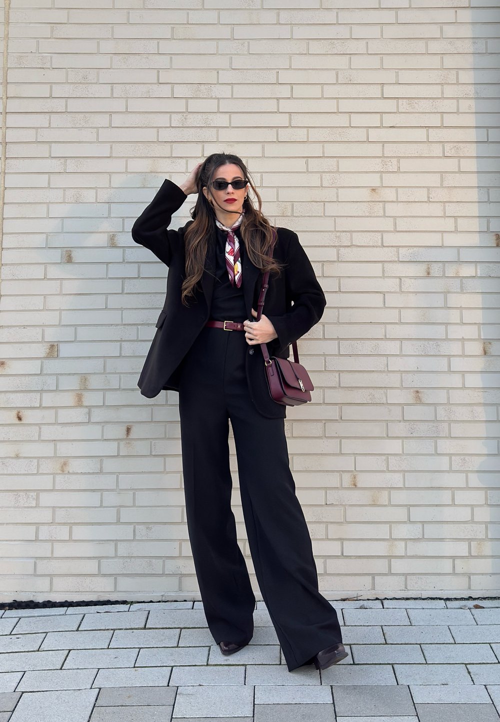 Femme en costume noir avec ceinture et sac bordeaux, foulard à fleurs, lunettes de soleil, debout contre un mur en briques claires, tenant ses cheveux d'une main.