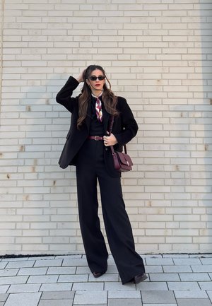 Mujer con traje negro, cinturón y bolso burdeos, pañuelo floral, gafas de sol, de pie contra una pared de ladrillos claros, sujetándose el cabello con una mano.