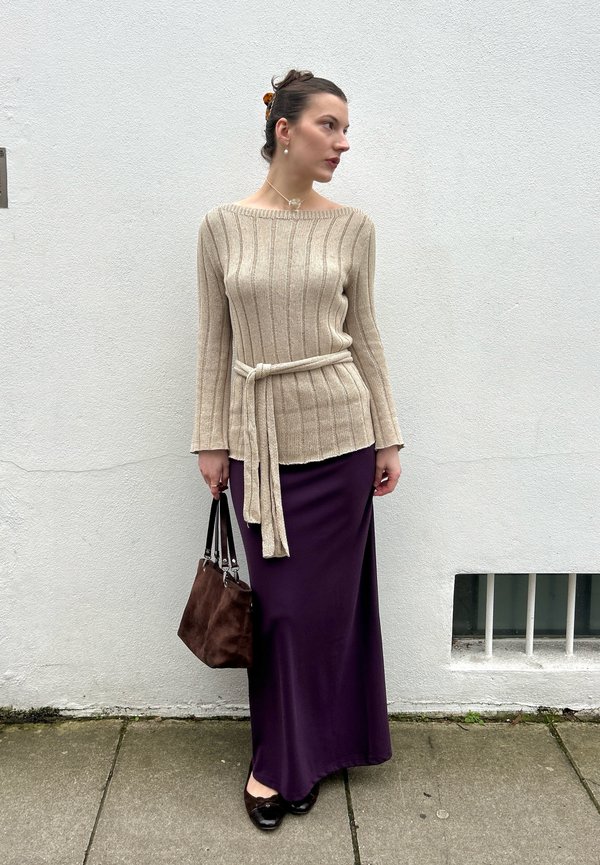 Donna che indossa un maglione beige a coste con cintura e una lunga gonna viola, che tiene una borsa marrone, in piedi contro un muro bianco.