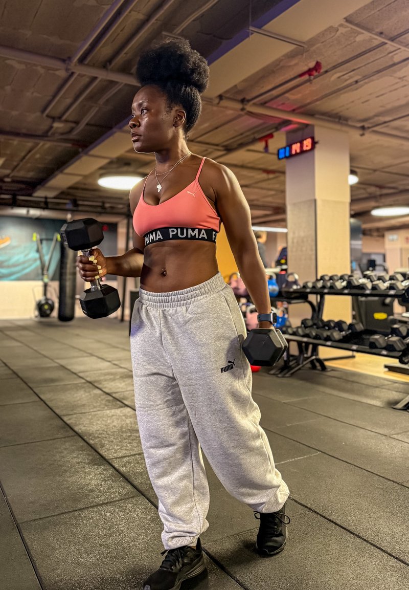 Kobieta na siłowni w sportowym biustonoszu i spodniach dresowych trzyma hantle, przygotowując się do treningu siłowego.
