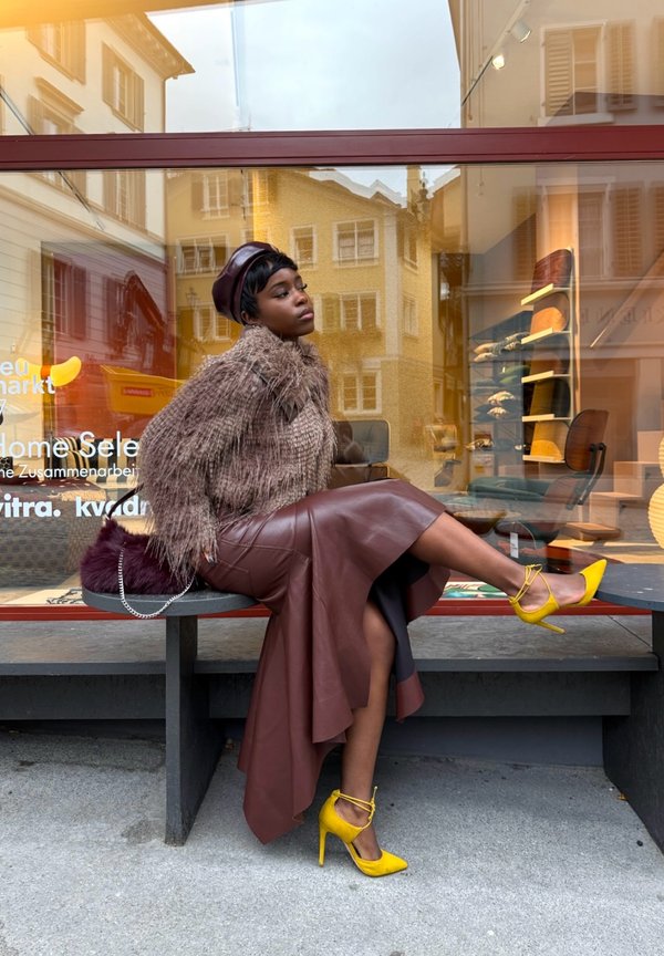 Brązowa skórzana spódnica midi z wysokim rozcięciem, puchaty brązowy sweter, żółte buty na wysokim obcasie i ciemny beret, siedząca na szarej ławce.