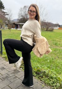 Beige strikket sweater, sorte udstående bukser, hvide sneakers. Holder en tan farvet plysjakke med lommer og står på en belagt sti omgivet af græs.