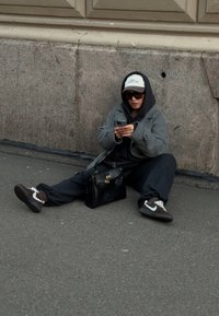 Személy szürke kabátban és fekete kapucnis pulóverben a járdán ül, okostelefont használ. Mellette fekete kézitáska, barna és fehér tornacipők.