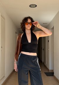 Černý halter top, černé sportovní kalhoty s bílými pruhy, hnědá kožená taška a oranžové sluneční brýle; kudrnaté vlasy; vnitřn�í prostředí s béžovými dlaždicemi.
