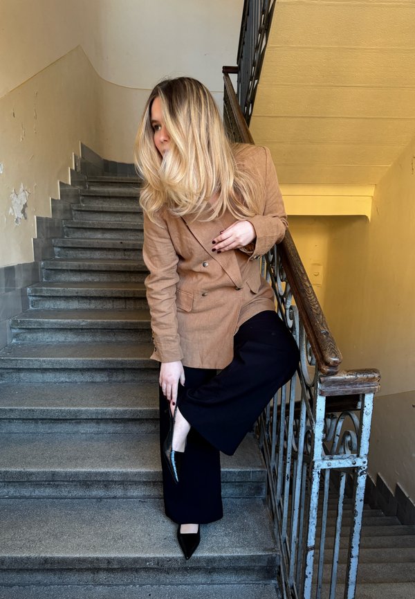Femme blonde en blazer marron et pantalon noir debout sur le palier d'escalier, ajustant une chaussure noire, avec une rambarde métallique et un mur usé en arrière-plan.