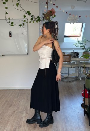 Mujer con tatuaje de pie en el interior con top blanco sin tirantes, falda negra, botas negras, sosteniendo un bolso negro, rodeada de plantas y muebles de comedor.