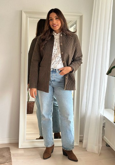 Mujer de pie en interior vistiendo una chaqueta acolchada marrón, blusa blanca de encaje, vaqueros azul claro y botines marrones frente a un espejo de cuerpo entero.