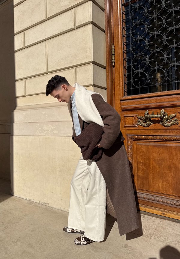Jonge man in een lange bruine jas, witte broek en schoenen met patroon, staand bij een sierlijke houten deur met een stenen muur op de achtergrond.
