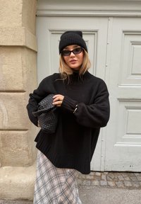Pull noir en maille surdimensionné à col haut, associé à une jupe à carreaux et un sac à main noir tissé. Les accessoires comprennent des lunettes de soleil foncées et un bonnet.