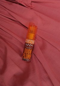 NYX Duck Plump lip produkt v čírej fľaši s oranžovým čerpadlom a výrazným oranžovým textom na jemnom ružovom textilnom pozadí.