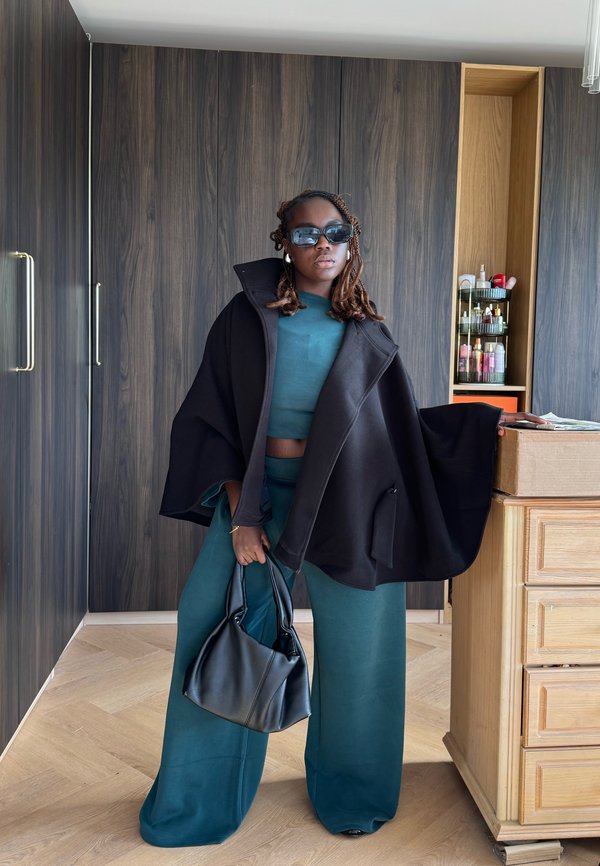 Mujer con el cabello trenzado con capa negra y conjunto azul verdoso, sosteniendo una bolsa negra, de pie en una habitación moderna con armarios y estantes de madera.
