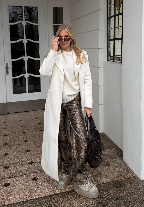 Vrouw in een lange witte jas, crème trui, slangenprint broek, grijze laarzen, met een zwarte tas met franjes, die haar zonnebril bij de ingang van een wit gebouw rechtzet.