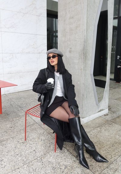 Mujer con abrigo negro, boina, gafas de sol, guantes y botas, sentada en una silla roja sosteniendo una taza de café afuera, cerca de una pared de cemento y una ventana.