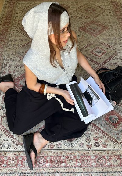 Sudadera gris sin mangas con capucha, pantalones sueltos negros y zapatillas negras. Sosteniendo un libro de fotos sobre una alfombra con patrones.