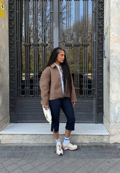 Mujer con el pelo largo trenzado, vestida con una chaqueta marrón, pantalones negros recortados, zapatillas blancas y sosteniendo un bolso blanco, de pie junto a una puerta metálica negra.