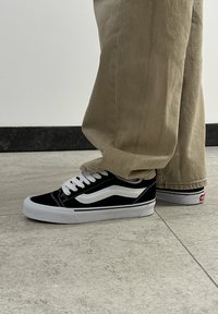 Μαύρα και λευκά αθλητικά παπούτσια με σουέτ και καμβά επάνω μέρος, διαθέτοντας παχιά λευκή σόλα και κλασικό σχέδιο κυμάτων στα πλάγια.