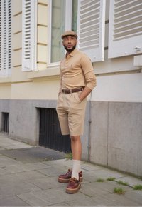 Béžová polokošile s dlouhým rukávem, khaki kraťasy, hnědý kožený opasek, hnědé platformové boty a bílé ponožky. Pozadí tvoří neutrální budova s okenicemi.